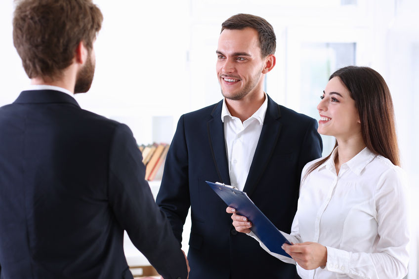 Three people in business attire are having a conversation; a man shakes hands with another while a woman, acting as a BSL interpreter and holding a clipboard, stands beside them, smiling.