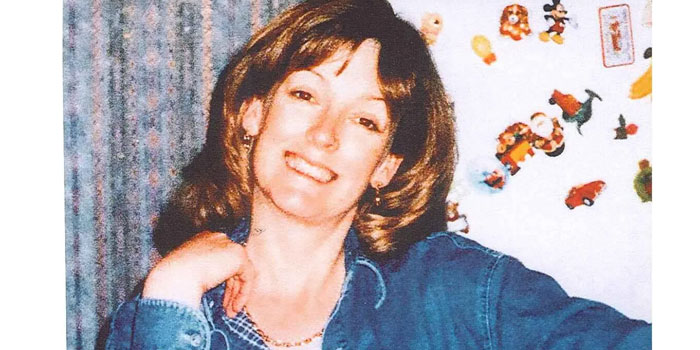 A woman with shoulder-length hair smiles at the camera, wearing a blue shirt. The background shows a curtain and a wall decorated with colorful magnets or stickers—perfect for practicing forensic lip reading skills.