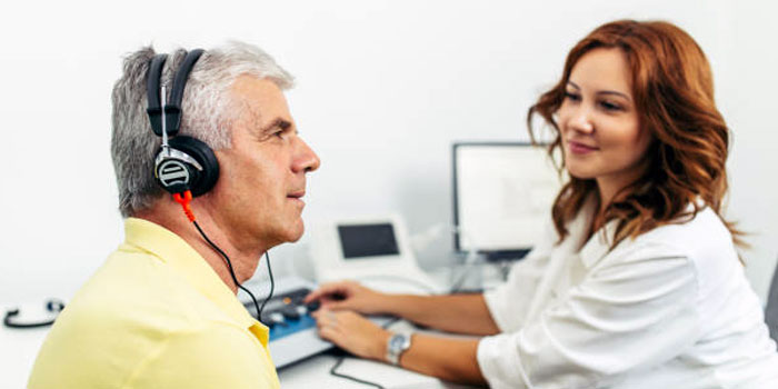 An older man wearing headphones undergoes a hearing test with an audiologist in a clinical setting.