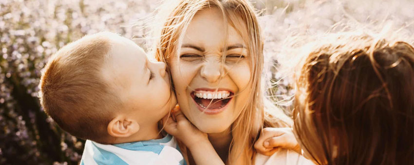 A woman smiles with her eyes closed as two children embrace her outdoors in bright sunlight, sharing a moment of warmth and connection through BSL.