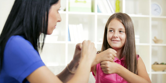 An adult and a child sit facing each other, using sign language to communicate in a bright room with shelves in the background, while a BSL interpreter supports their conversation.