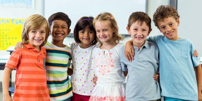 Six children stand closely together in a classroom during a BSL lesson, smiling at the camera. Some have their arms around each other. The background shows part of a whiteboard and a yellow poster.