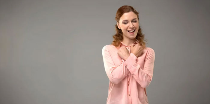 A woman with curly hair in a pink blouse stands against a gray background, smiling with her arms crossed over her chest as she uses BSL.
