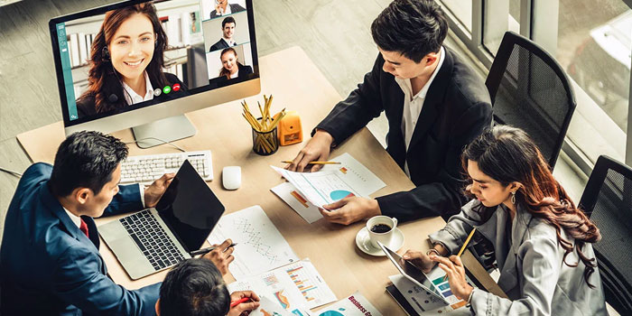 Four people in business attire sit at a table with charts and a laptop, participating in a video conference with colleagues shown on a computer screen—ideal for business captions.