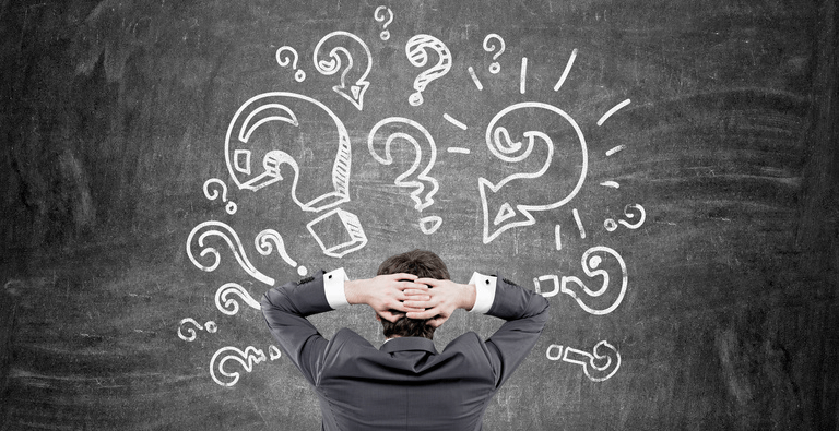 A person in a suit stands facing a chalkboard filled with large drawn question marks, hands resting on the back of their head—perhaps pondering the challenges that captioning companies help solve.