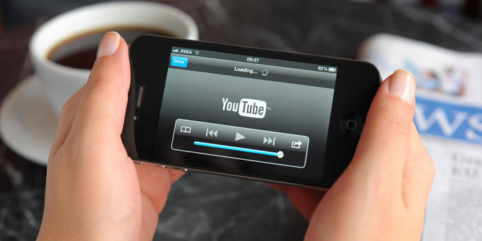 Person holding a smartphone displaying the YouTube loading screen, ready to caption online videos, with a cup of coffee and a newspaper in the background.