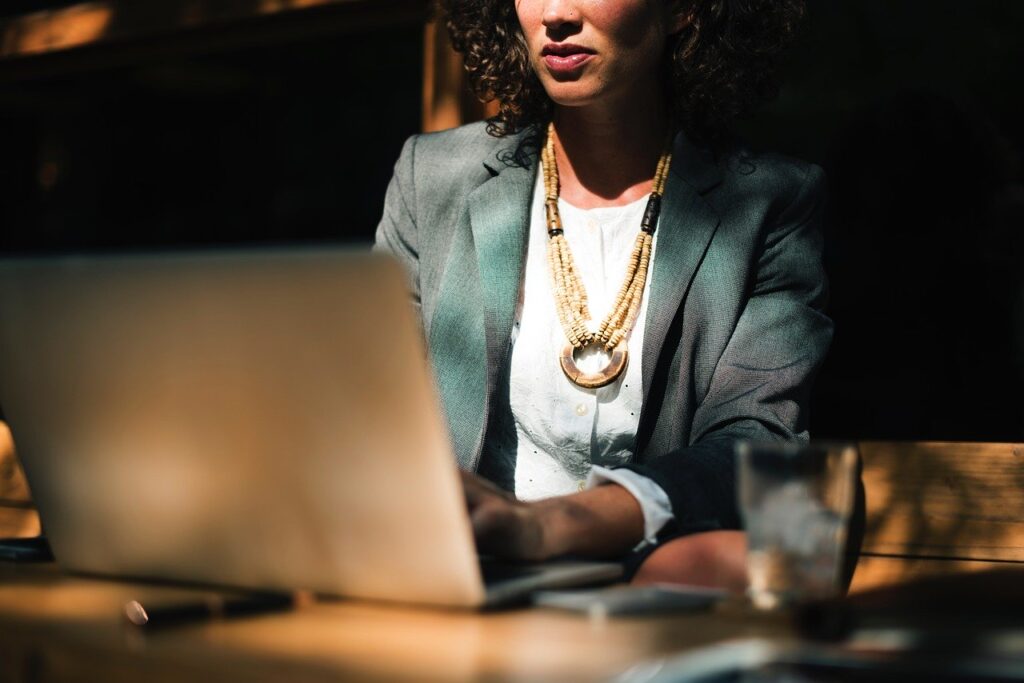 A woman in a blazer sits at a table working on a laptop, with a glass and notebook nearby.