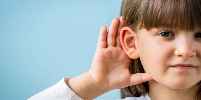 A young girl with brown hair holds her hand to her ear, appearing to listen closely—perhaps experiencing the effects of hearing loss—against a light blue background.