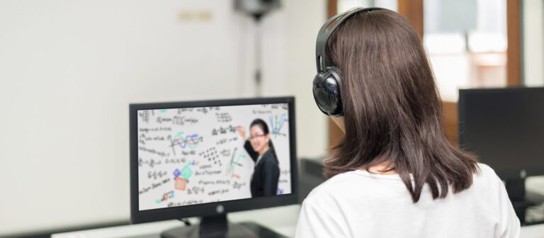 A person wearing headphones sits at a desk and watches an online lesson with diagrams, a teacher, and live captioning for education displayed on a computer monitor.