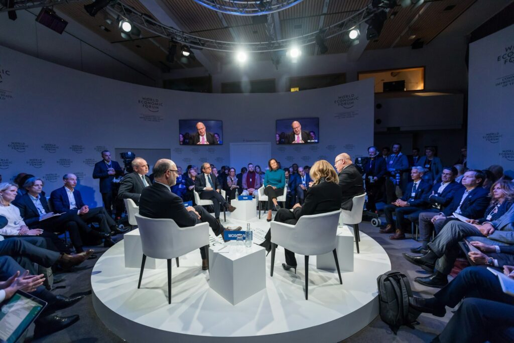 People sit in a circular arrangement on stage for a panel discussion, with an audience and cameras surrounding them in a conference room supported by remote captioning services.