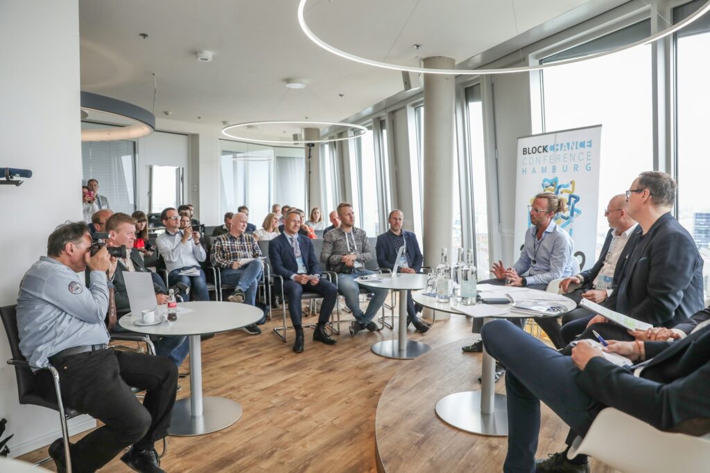A group of people attend a panel discussion about the partner program in a modern conference room with large windows at a blockchain conference in Hamburg.