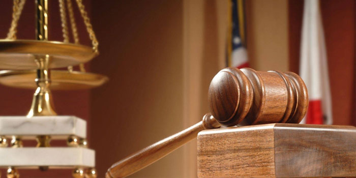 A wooden gavel rests on a block in a courtroom, with the scales of justice and flags visible in the background, as a forensic lip reader prepares to analyze silent courtroom footage.