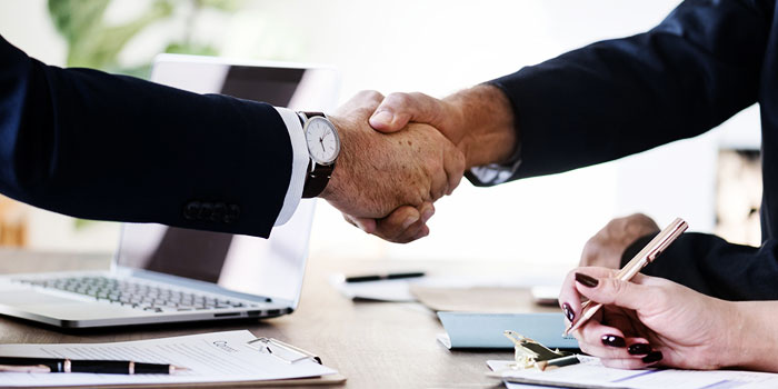 Two people in business attire shaking hands across a desk, with paperwork, a pen, and a laptop visible in the background—an ideal setting to hire a stenographer for your important meetings.