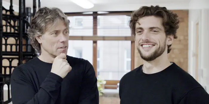 Two men wearing black shirts stand indoors; one looks thoughtful with his hand on his chin while the other smiles, as they enjoy learning BSL together.