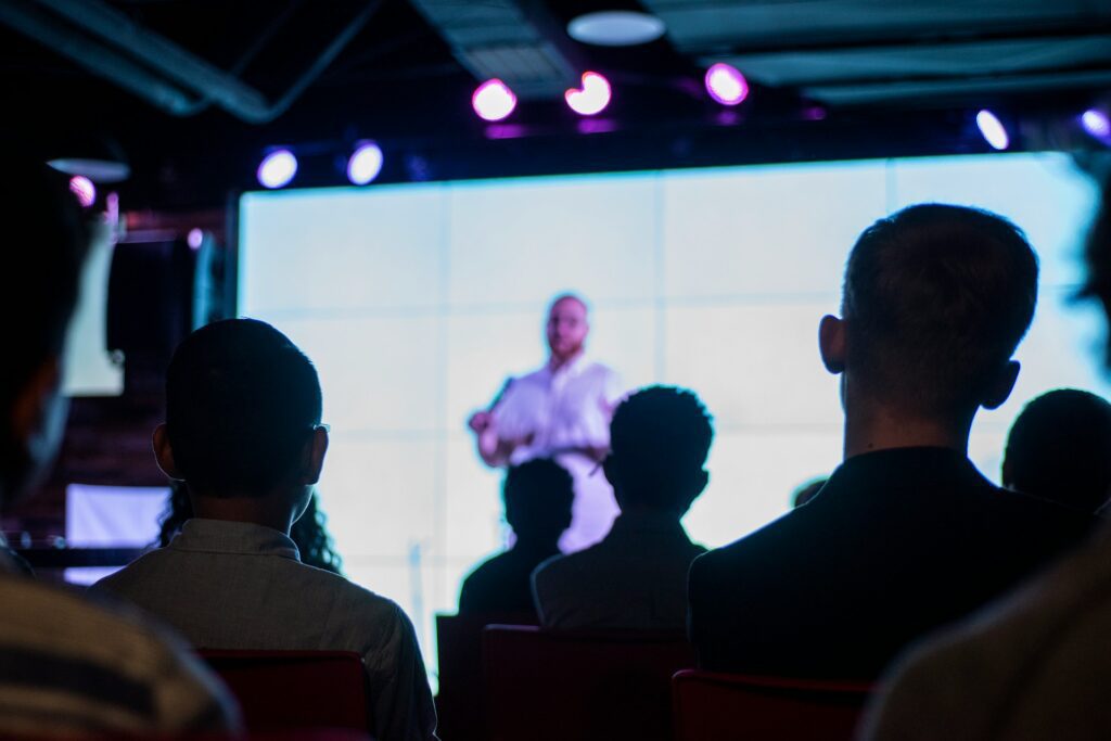 Audience members sit facing a stage where a person stands in front of a large, brightly lit screen, giving a presentation supported by remote captioning services for enhanced accessibility.