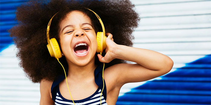 A child with curly hair wearing yellow headphones is singing or shouting joyfully in front of a blue and white striped background—showing how you can protect your child’s hearing while they have fun.