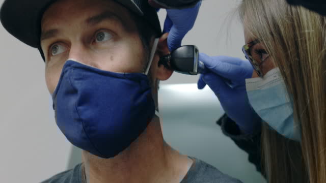 A healthcare professional wearing gloves and a mask examines a man's ear with an otoscope, checking for signs of hearing loss; the man is also wearing a face mask and a cap.