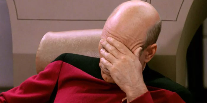 A person in a red and black uniform sits in a chair with their hand covering their face, appearing frustrated or exasperated, as if they feel like they're talking to a deaf person.