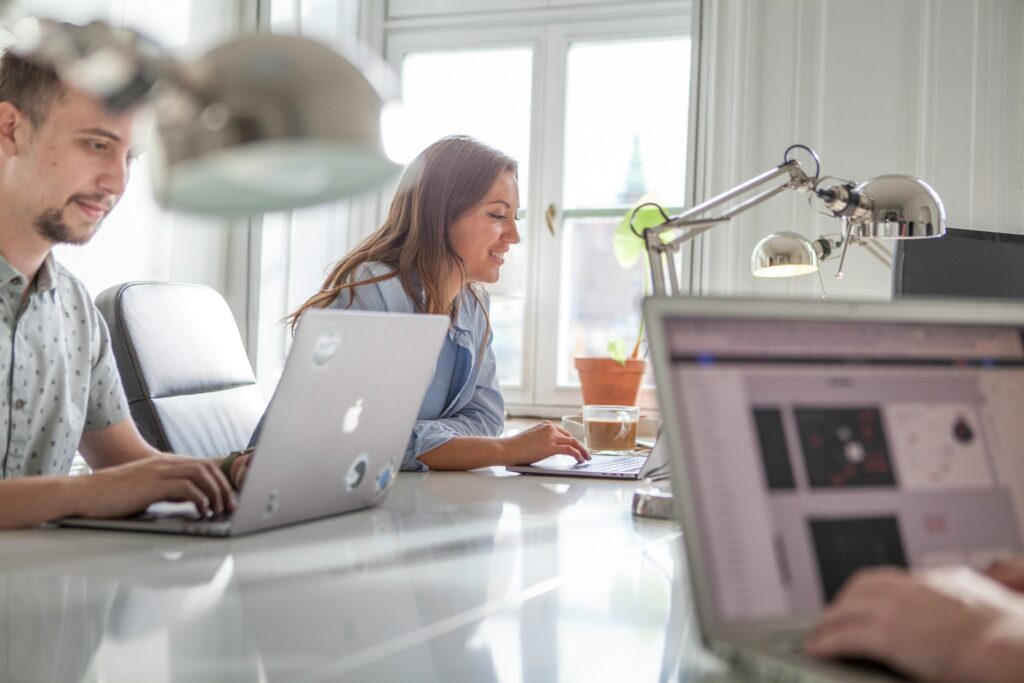 Three people work at a bright office desk with laptops and desk lamps, sharing testimonials about their projects. A window and potted plant are visible in the background.