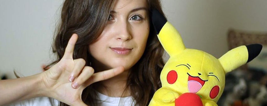 A person with long brown hair, like Rikki Poynter, holds a Pikachu plush toy and forms the “I love you” sign in American Sign Language with their hand.