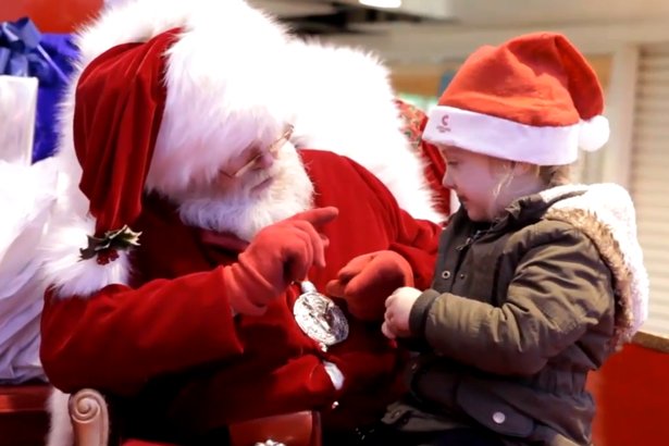 Santa Claus sits with a young child wearing a Santa hat, smiling and pointing at each other in a festive, Christmas deaf friendly setting.