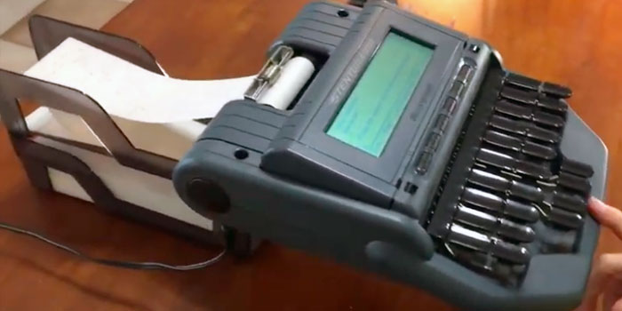 A modern stenograph machine, essential for any skilled stenographer, features a digital display and connected paper roll as it sits on a wooden surface.