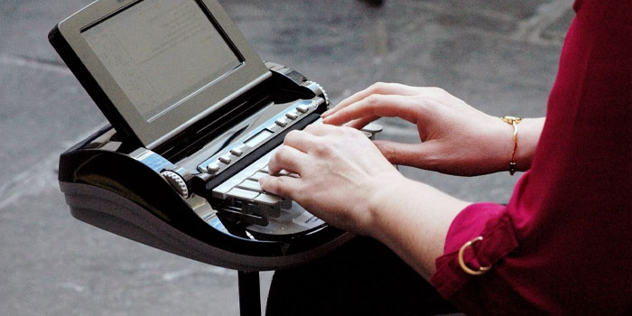 A stenographer in a red shirt types on a stenography machine while watching text appear on the screen.