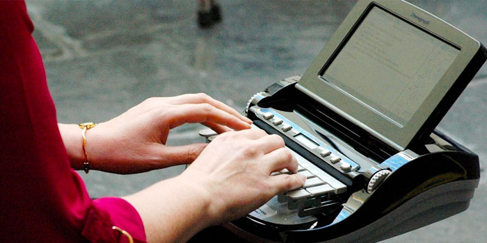 A stenographer in a red sleeve types on a stenograph machine with an attached display screen.