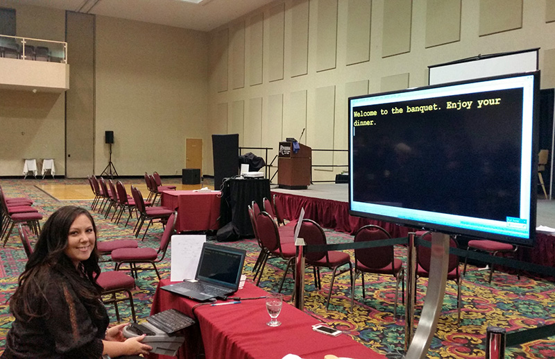 A woman sits at a laptop in a conference room with a large screen displaying live captioning and the text, "Welcome to the banquet. Enjoy your dinner." Red chairs are arranged facing a stage.