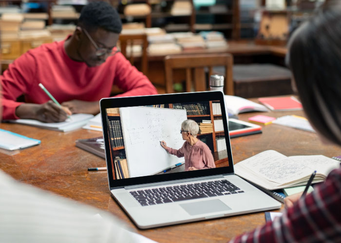 A laptop on a table displays a person teaching at a whiteboard with live captioning. Two people sit nearby, taking notes and studying with open books and notebooks.