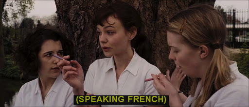 Three young women in white shirts sit by a tree, two holding cigarettes, with yellow subtitles displayed at the bottom reading "(SPEAKING FRENCH)".