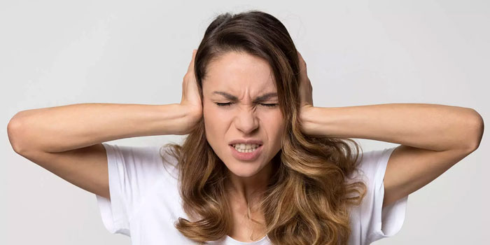 Woman with long hair in a white shirt covers her ears with her hands and closes her eyes tightly, appearing to be in discomfort, possibly due to tinnitus or being disturbed by noise.