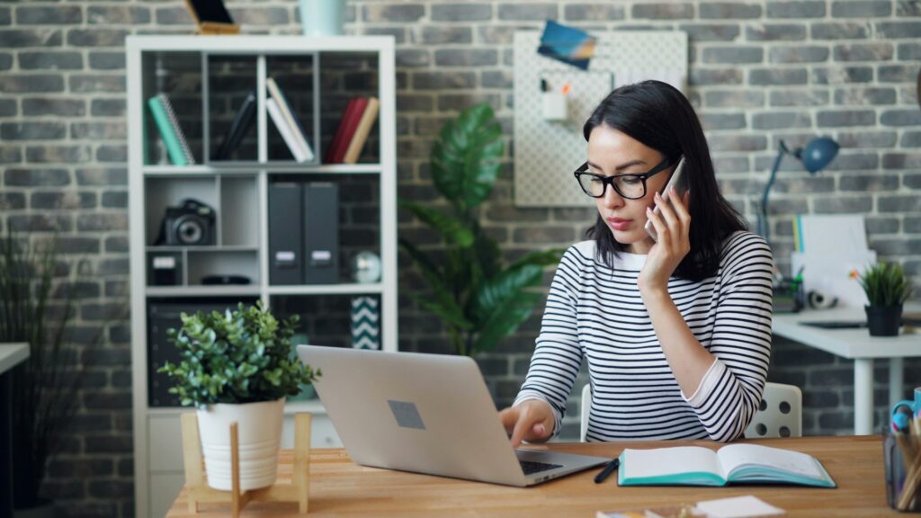 Woman wearing glasses works on a laptop at a desk, talking on a cellphone, with notebooks and a potted plant in a home office setting—perfect for remote work or using live captioning services.