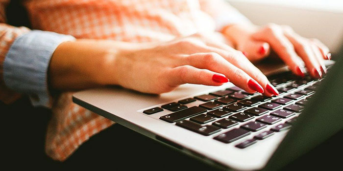 Close-up of a person with red nail polish typing on a laptop keyboard, wearing an orange and white checkered shirt, working on offline captioning.