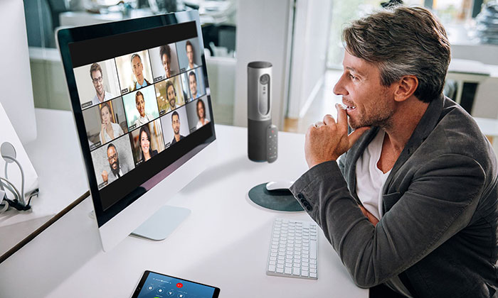 A man sitting at a desk participates in a video conference call with multiple people displayed on a computer screen, using live captions to follow the conversation clearly.