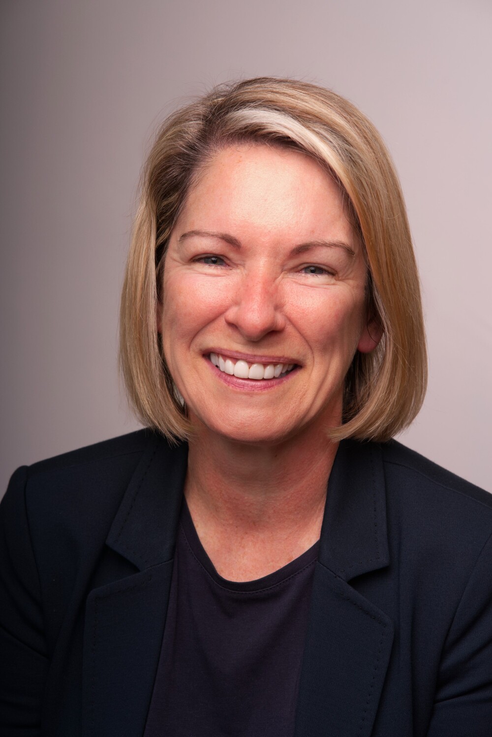Tina Lannin, a woman with short blonde hair, is smiling while wearing a dark blazer and top, pictured against a neutral background.