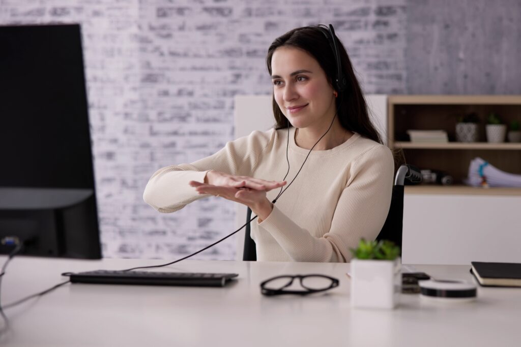 A woman wearing headphones sits at a desk in an office, using British Sign Language Interpreting in front of a computer monitor.
