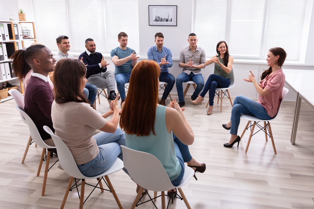 A group of people sit in a circle on chairs in a bright room, participating in a deaf awareness training course, with one woman speaking and others listening and gesturing.